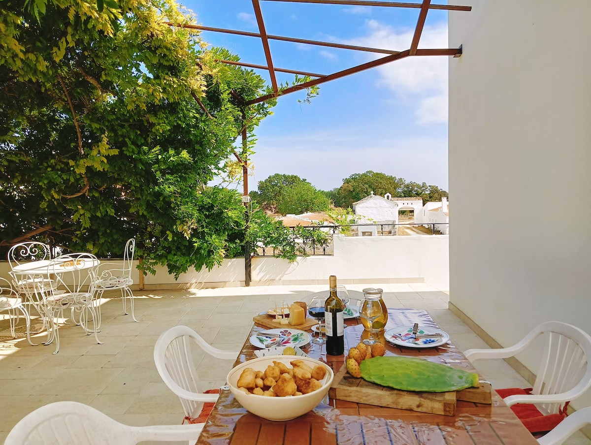 An outdoor dining area features a wooden table set with a variety of foods, including wine and freshly baked items. Nearby, a white wrought-iron table and chairs are visible, surrounded by greenery. The sky above is bright with soft clouds.