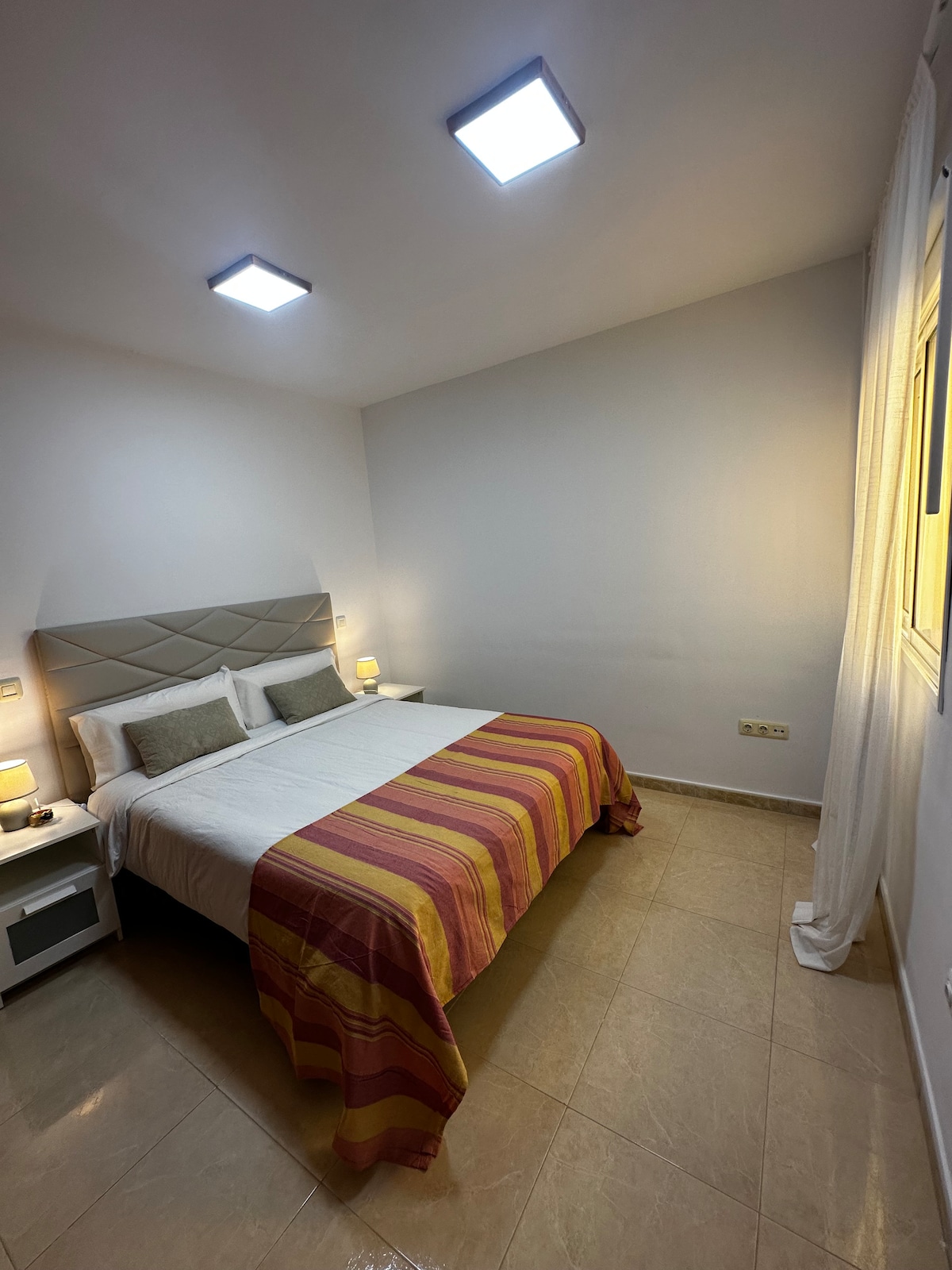 A well-lit bedroom features a spacious bed adorned with a striped blanket. Soft bedside lamps illuminate the nightstands on either side. A window allows natural light to enter, complemented by light-colored curtains, and the walls are finished in a neutral tone.