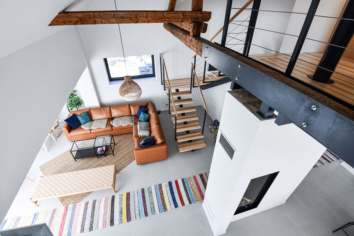 An open living area is highlighted by a modern orange sofa, arranged around a glass-top coffee table. A staircase with wooden steps is visible, leading to a lofted section. A colorful patterned rug runs across the tiled floor, enhancing the airy space.