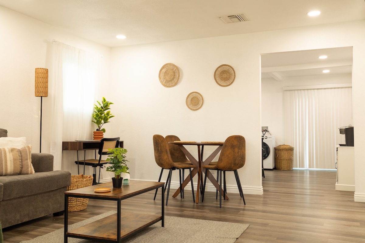 A spacious living area features a comfortable gray sofa and a wooden coffee table, adorned with a small plant. A dining table with four chairs occupies the background, surrounded by decorative wall art. Natural light filters through sheer curtains, enhancing the open, inviting space.