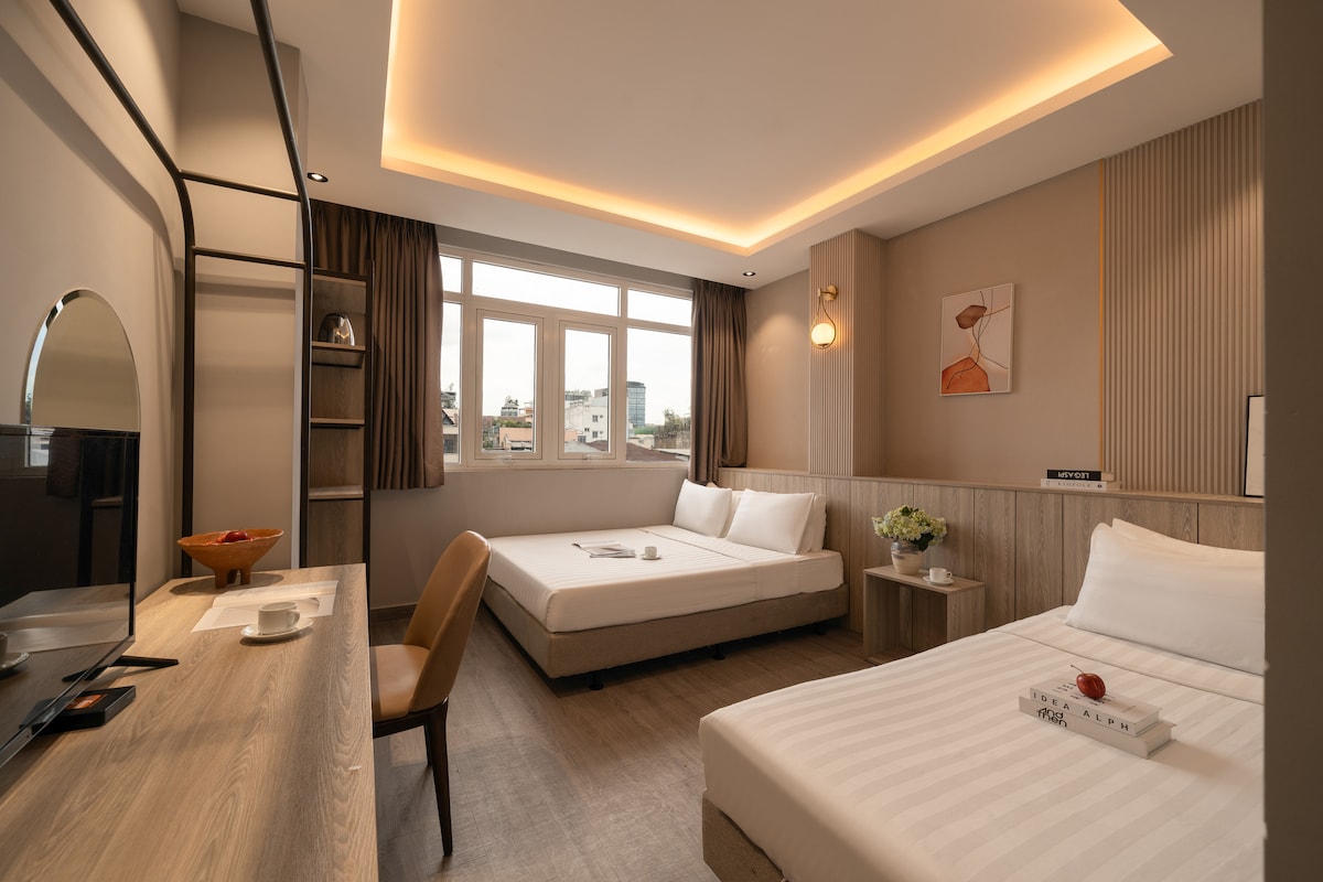 A spacious room is shown with a well-designed layout, featuring a double bed and a single bed. Natural light enters through large windows, illuminating the neutral tones of the decor. A small table with a flower arrangement and a chair is positioned near the window, while a television is mounted on the wall.