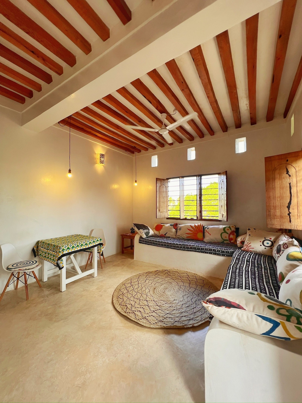 A spacious living area is presented with local art fabrics and comfortable seating arrangements. Natural light enters through window openings, illuminating the high ceiling adorned with wooden beams. A circular rug anchors the space, complemented by a dining table set for conversation.