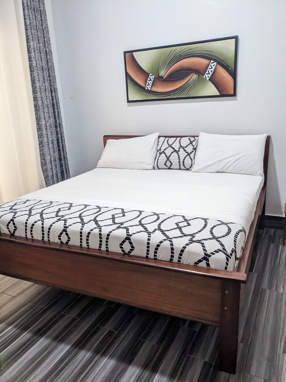 A clean sleeping area features a spacious bed with a patterned blanket and three pillows. A piece of abstract wall art adds a touch of style above the headboard. Natural light enters through sheer curtains, illuminating the wooden floor.