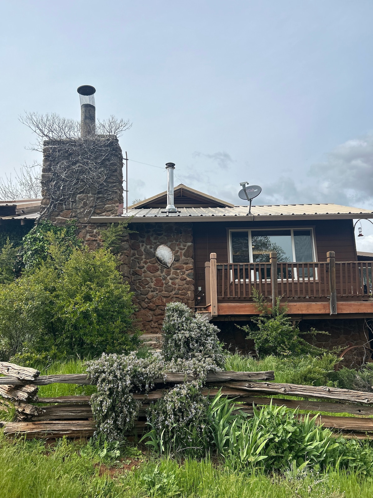 A rustic cabin is set against a natural backdrop, featuring a stone chimney and wooden balcony. Greenery surrounds the structure, with wildflowers peeking through. The cabin's earthy tones blend harmoniously with the landscape, while large windows reveal the inviting interior.