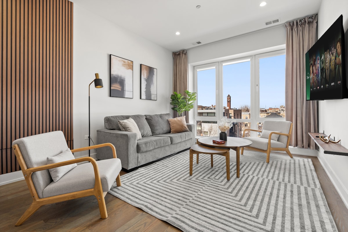 A modern living area features a gray sofa with accent chairs set beside large windows, allowing natural light. A patterned area rug covers the floor, complemented by a minimalist coffee table. Wall art adds subtle elegance, enhancing the overall inviting layout.