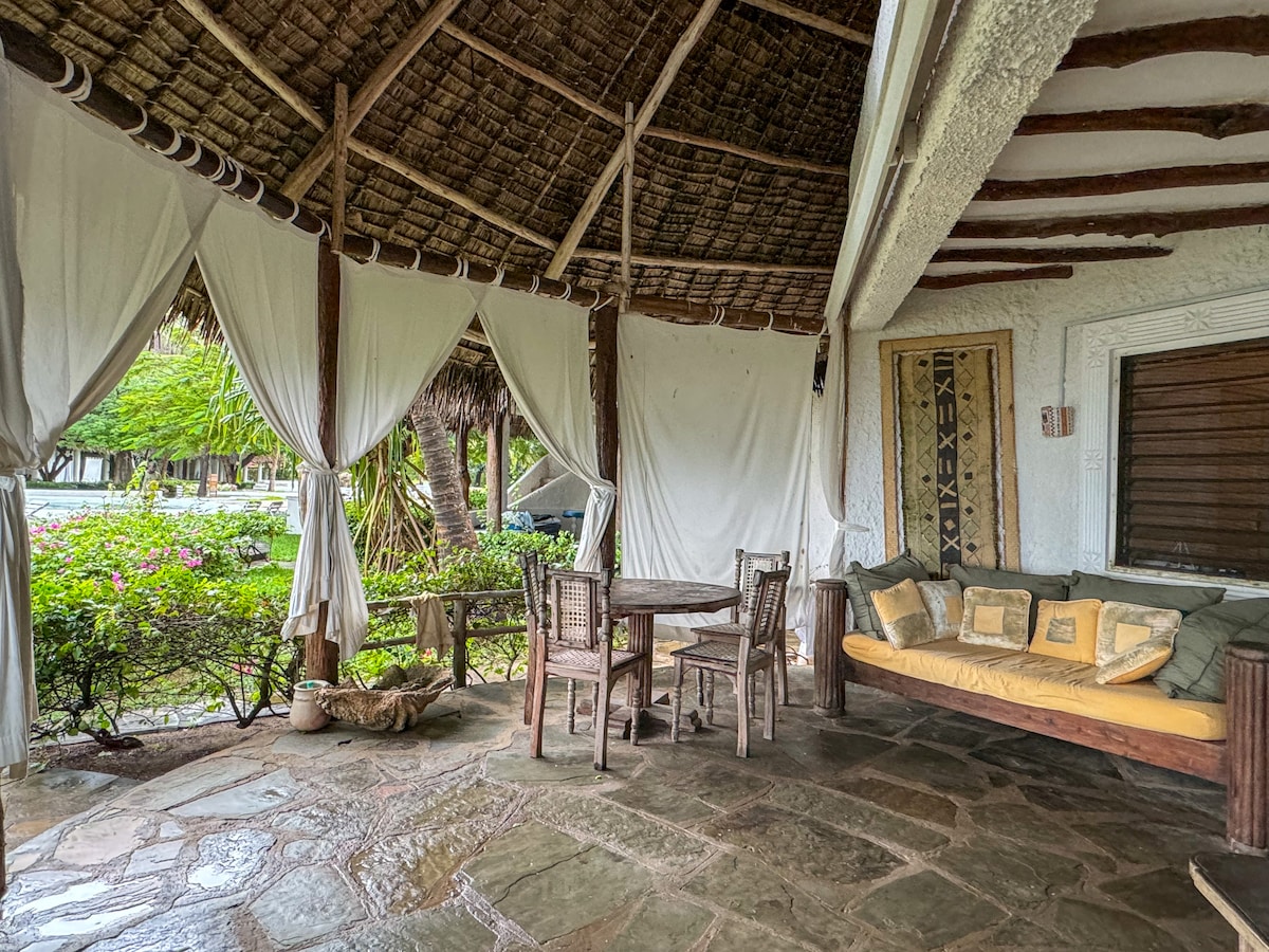 A covered terrace features a rustic dining table set for four, accompanied by a plush seating area with cushions. Textured stone flooring contrasts with the surrounding greenery, while sheer white drapes provide a soft separation from the outdoors.