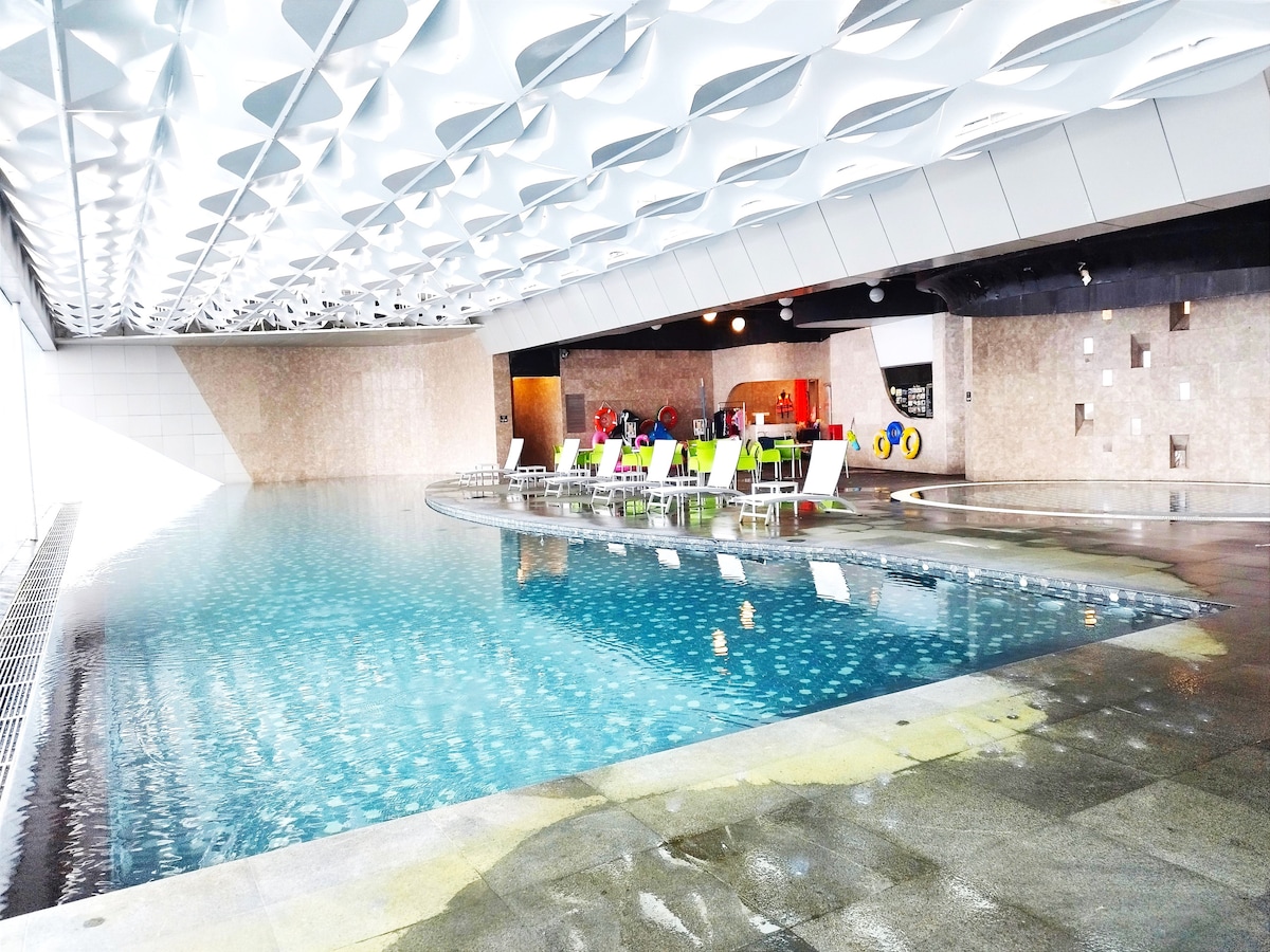 An indoor heated swimming pool is seen, featuring a smooth turquoise surface reflecting light from overhead fixtures. Surrounding the pool are white lounge chairs arranged neatly, providing a comfortable space for relaxation. The sleek design is enhanced by a modern ceiling with geometric patterns.