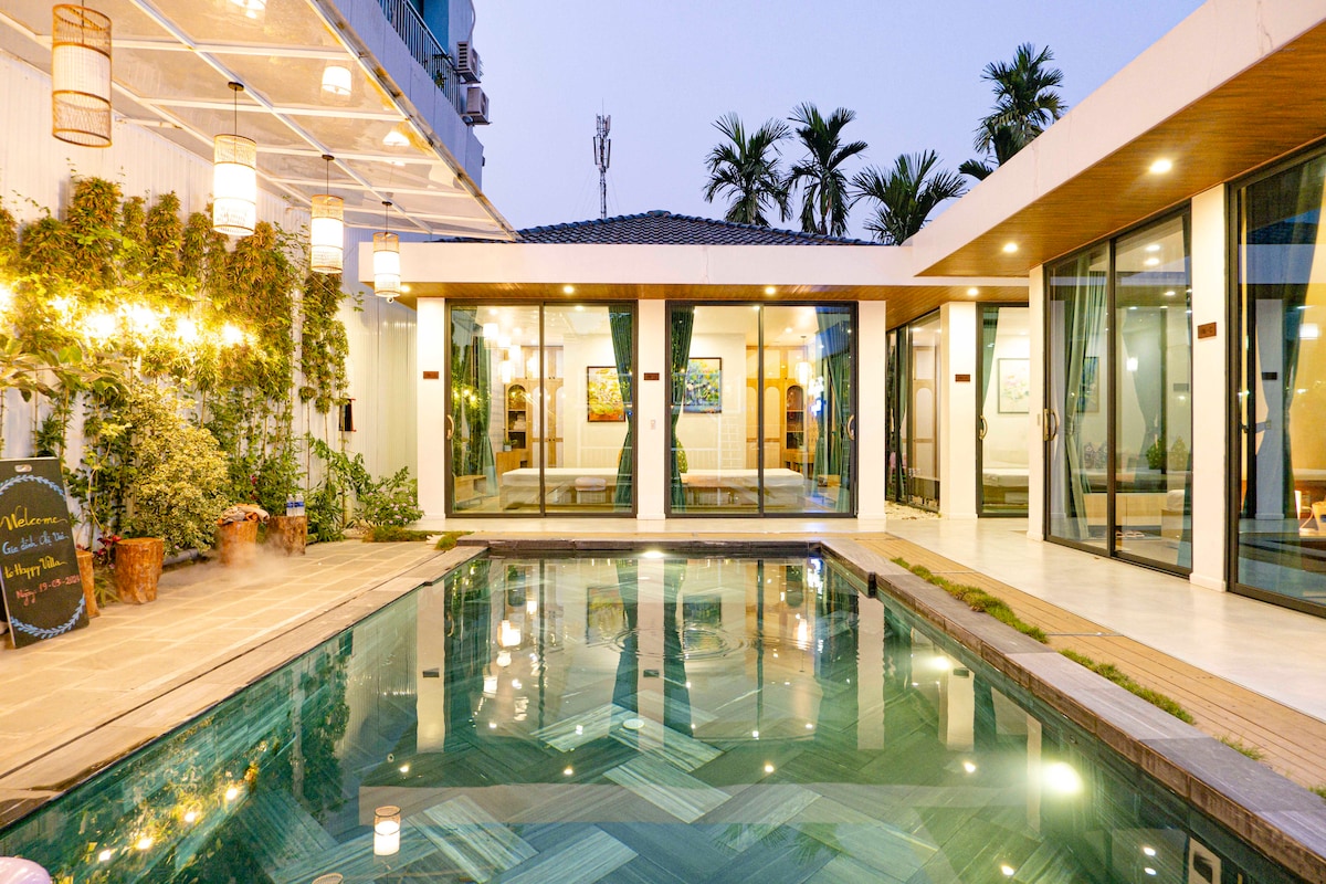 A spacious outdoor area features a clear swimming pool surrounded by tiled flooring. Large glass doors open into an inviting common area, framed by tropical plants and soft lighting. The setting is complemented by a serene evening atmosphere and palm trees in the background.