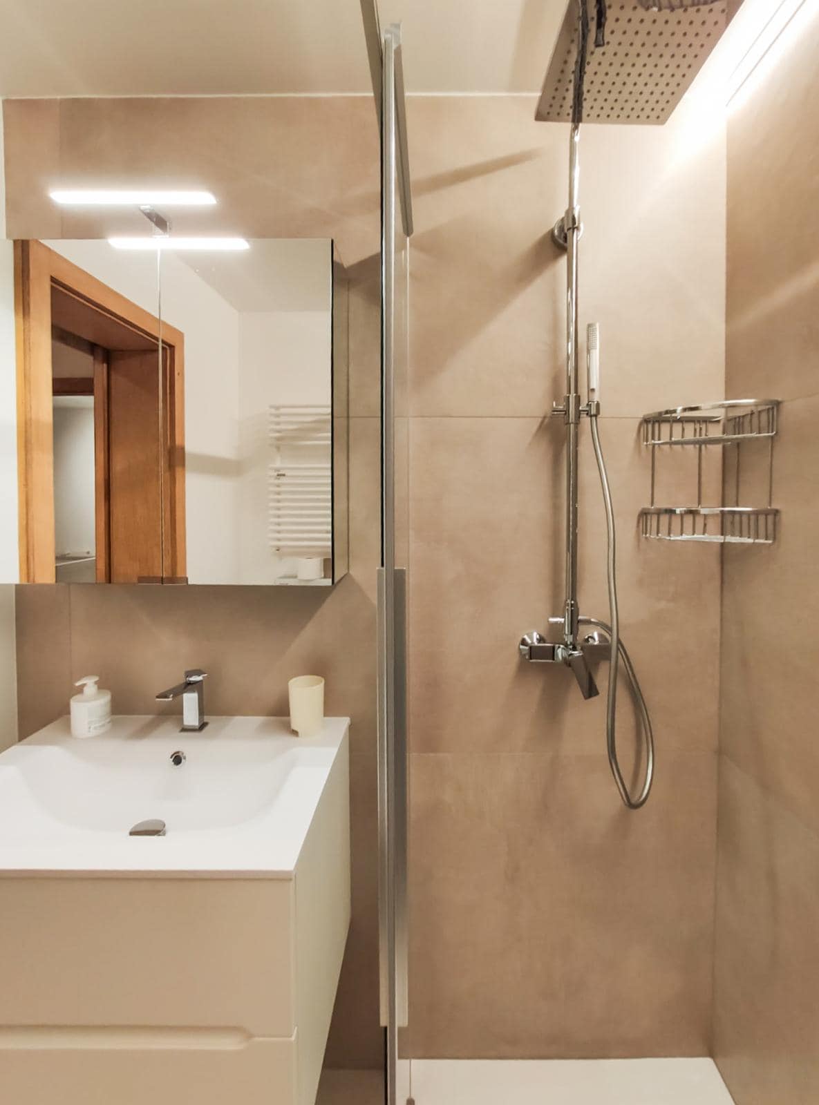 The modern bathroom features neutral-toned wall tiles and a walk-in shower with a glass door. A compact sink with an integrated cabinet is positioned beneath a well-lit mirror. Shelving is mounted for convenient storage of toiletries.
