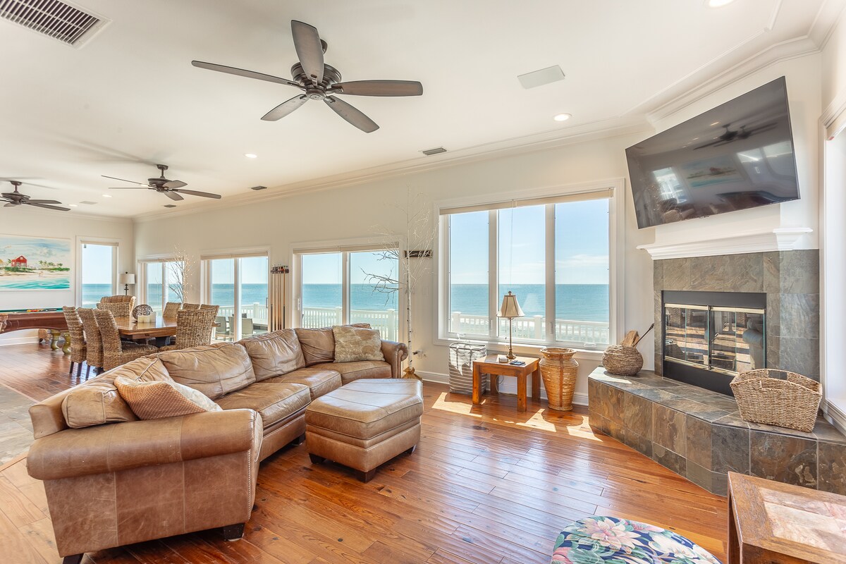 An open living area features large windows providing unobstructed views of the beachfront. A spacious sectional sofa, a stone fireplace, and a smart TV are prominently displayed. The area offers a warm, inviting space with hardwood floors and ceiling fans, enhancing comfort and convenience.