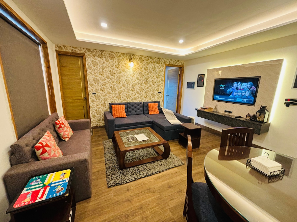 The living area features a sectional sofa with orange cushions, a coffee table, and a dining table that seats four. A 50-inch flat screen TV is mounted on the wall. Soft lighting highlights the floral wallpaper and wooden accents.