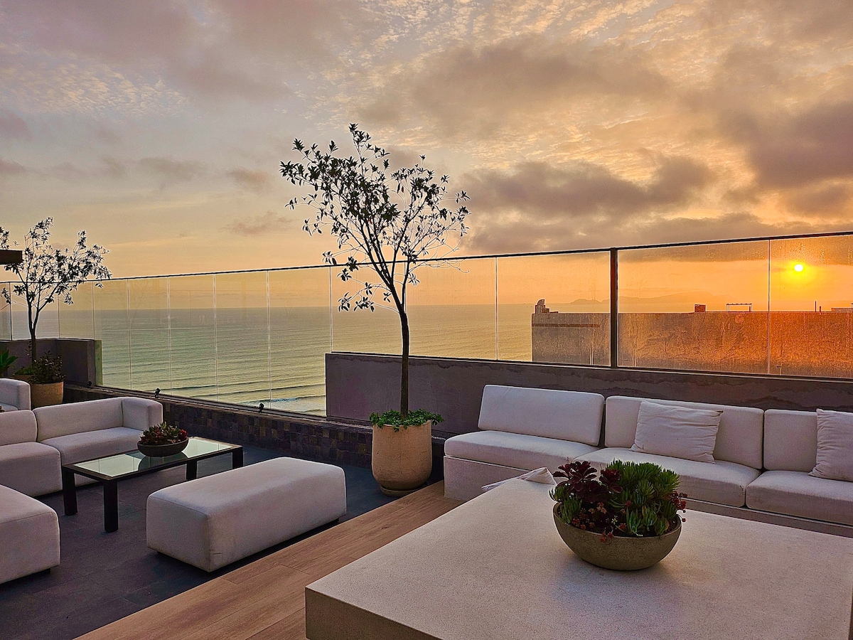 A rooftop lounge area features modern white seating arranged around a central table. Planters with greenery are placed nearby. A serene sunset illuminates the sky and reflects off the ocean, creating a peaceful atmosphere.