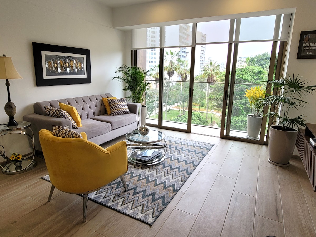 A bright living area features a gray sofa accented with yellow cushions, complemented by a round glass coffee table. A patterned area rug lies on the floor, while large glass doors provide a view of the greenery outside, enhancing the room's airy feel.