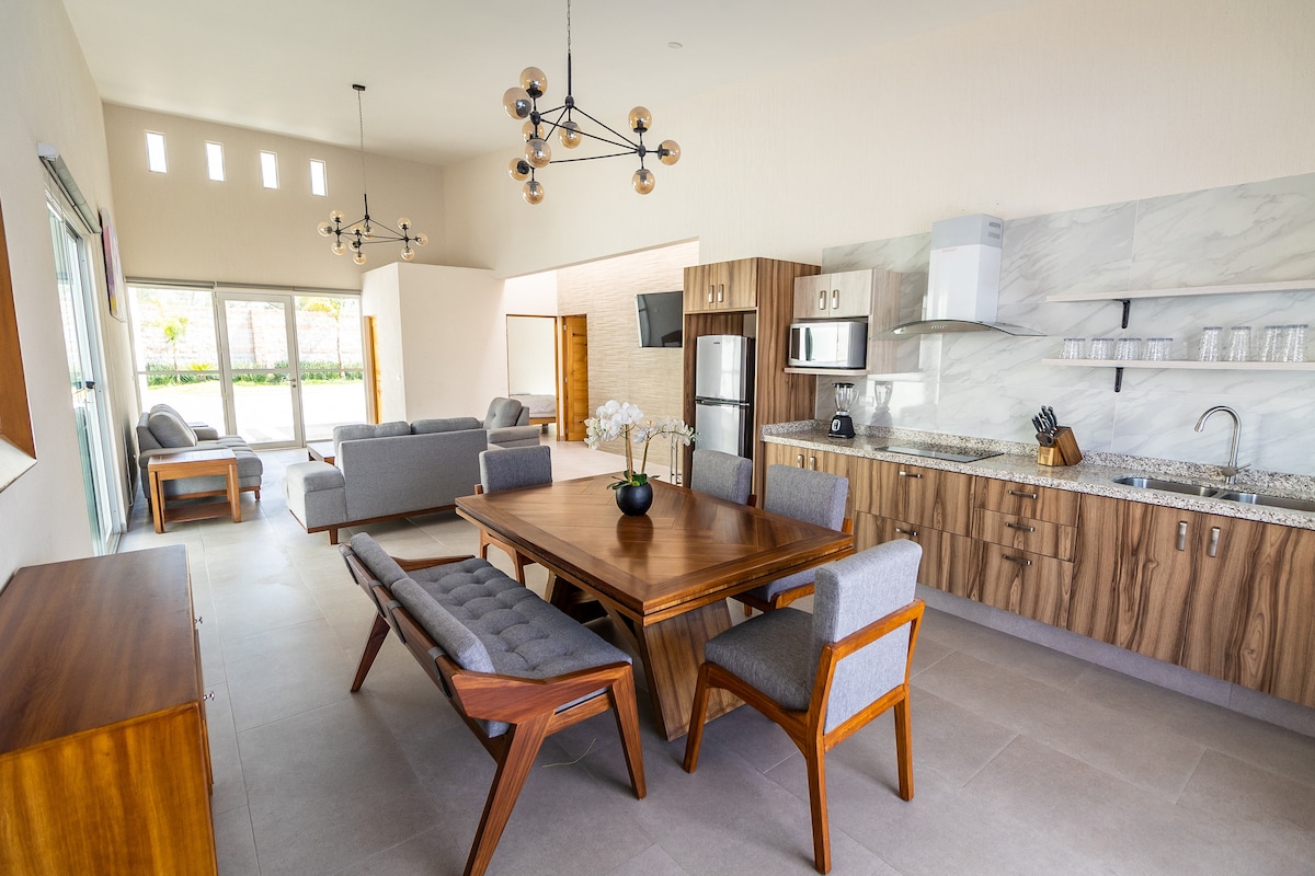An open-concept living and dining area features a wooden dining table surrounded by upholstered chairs. A modern kitchen is visible, showcasing stainless steel appliances and a marble backsplash. Comfortable seating is arranged nearby, with natural light filtering through large glass doors.