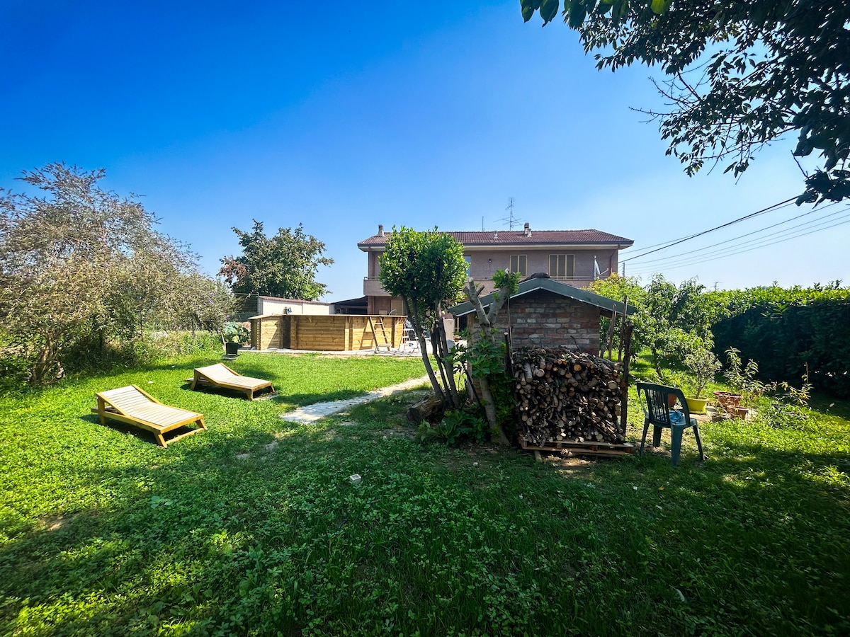 A spacious private garden is showcased, featuring sun loungers, a wood pile for the barbecue area, and a storage shed. The house stands in the background, surrounded by greenery and low shrubs, providing a relaxed outdoor environment.