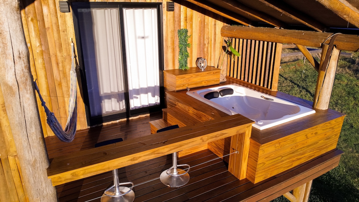 A wooden deck extends from the cabin, featuring a private jacuzzi with sleek wooden surrounds. Bar stools are positioned at a small counter, with a hammock hanging nearby. Large glass doors provide access to the interior, while natural light enhances the outdoor space.
