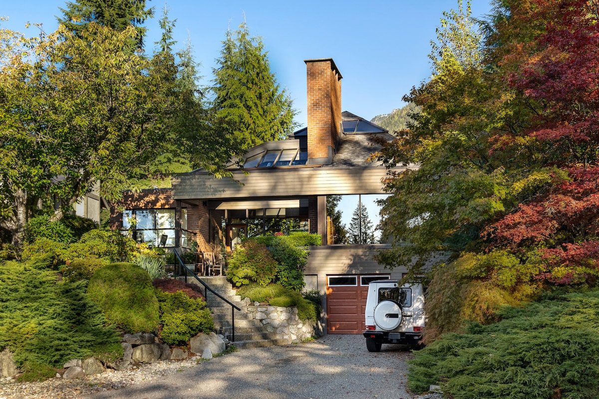 The exterior of a modern home is framed by lush greenery and colorful foliage. A large deck with seating overlooks the entrance, while a driveway provides access to an attached garage. The sloped roof and large windows enhance the contemporary design against a backdrop of towering trees.
