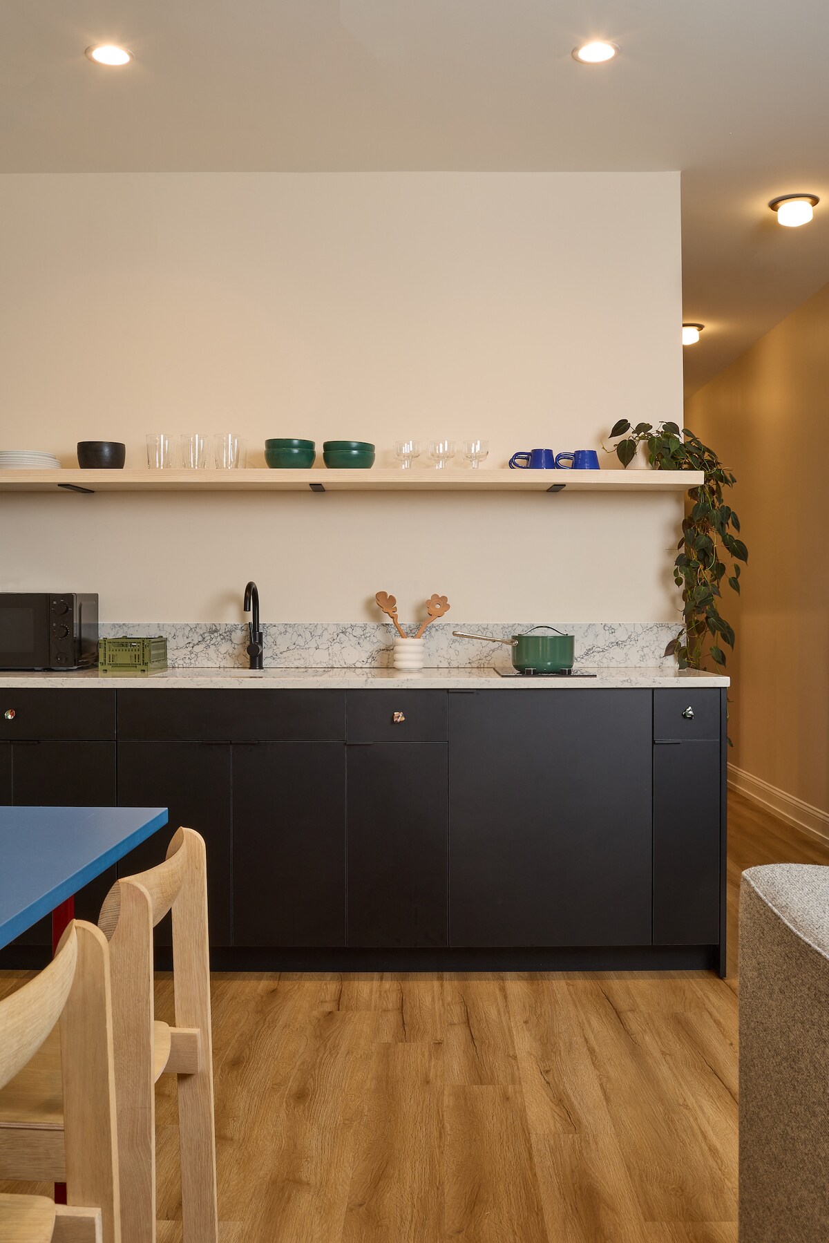 A sleek kitchen area is highlighted by dark cabinetry and a light marble countertop. Open shelving holds glassware and decorative items, while a small potted plant adds a touch of greenery. A compact microwave and sink complete the functional space, with warm lighting enhancing the overall appearance.