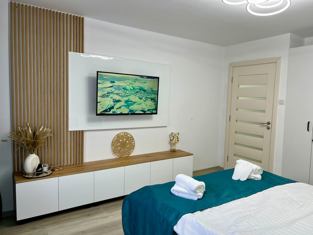 A modern bedroom is equipped with a Smart TV mounted on the wall, situated above a minimalist wooden console. Soft lighting complements the serene decor, featuring a decorative wall and neatly arranged towels on the bed. The space is designed for comfort and relaxation.
