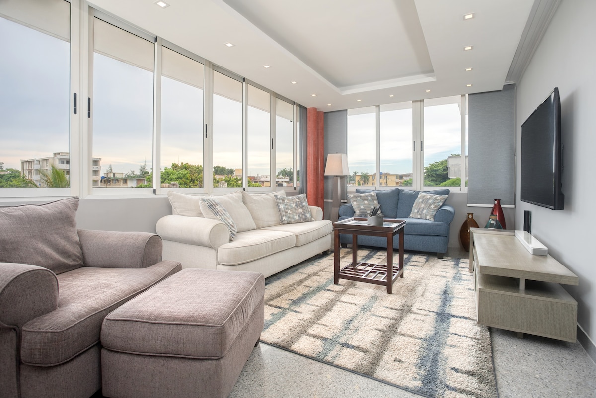 A modern living room features large windows allowing natural light to fill the space. A light-colored sofa, accent chair, and coffee table are arranged around a plush area rug. A television is mounted on the wall, offering an inviting spot for relaxation.