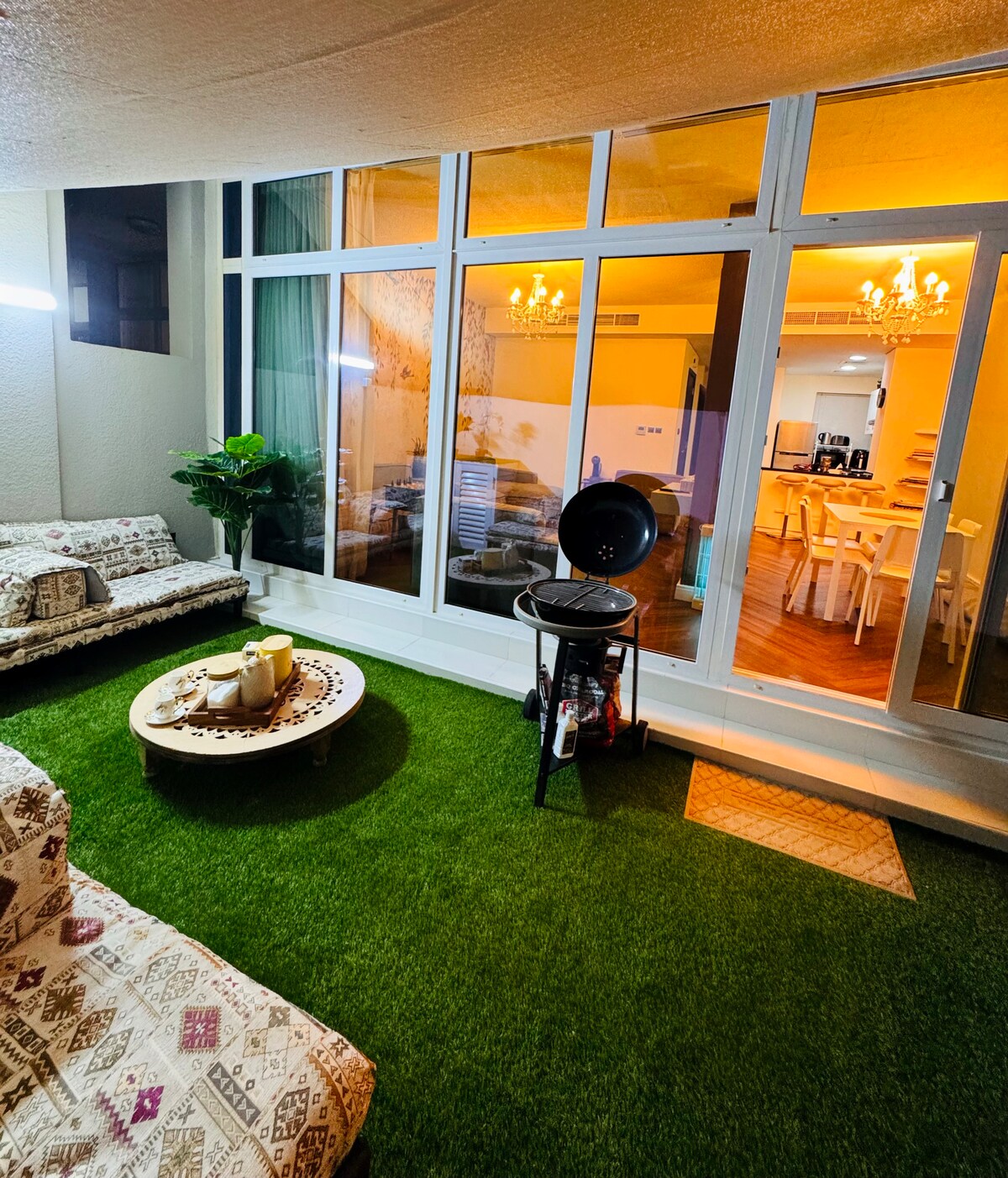 A cozy outdoor seating area is highlighted by artificial grass and comfortable sofas adorned with patterned cushions. A small coffee table is positioned at the center, displaying decorative items. Large windows allow natural light to fill the space, connecting it to the interior.