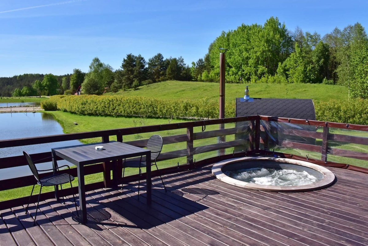 A wooden deck offers an inviting space with a round hot tub and a table for outdoor dining. Scenic views of lush greenery and a pond create a relaxing atmosphere. The surrounding area is tranquil, enhancing the natural landscape.