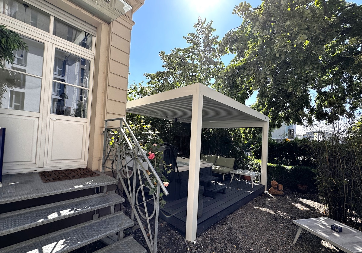 A covered outdoor seating area is presented, featuring a wooden deck and a dining table surrounded by modern chairs. Lush greenery and trees provide shade, while sunlight filters through, creating a bright and inviting space. The adjacent entrance showcases elegant stairs and detailed railings.