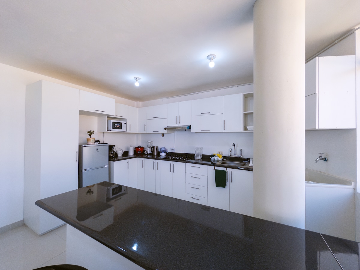 The kitchen features modern white cabinetry and sleek black countertops. A microwave, stove, and refrigerator are included among the appliances. Natural light illuminates the space, highlighting a central island with seating and ample room for meal preparation.