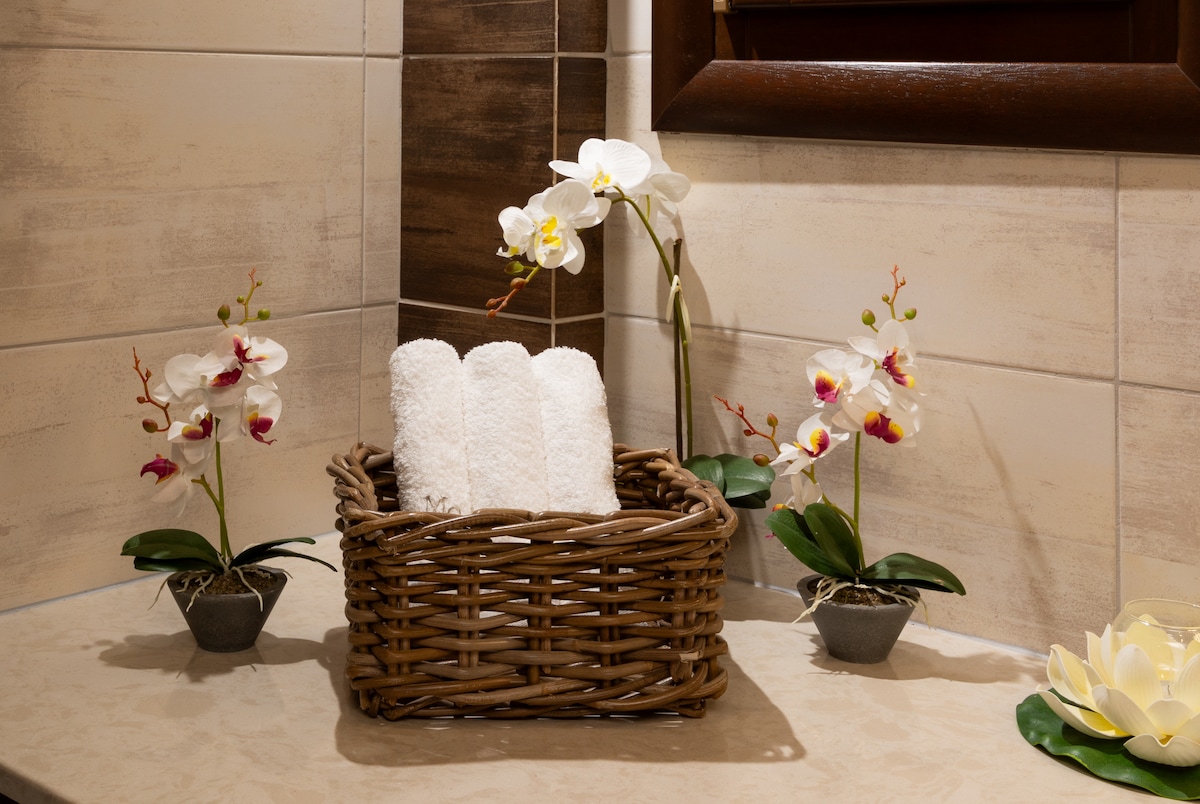 A wicker basket holds neatly rolled white towels, placed on a smooth countertop. Flanking the basket are potted orchids, adding a touch of nature. The neutral tiles provide a calming backdrop, enhancing the space's clean and organized appearance.