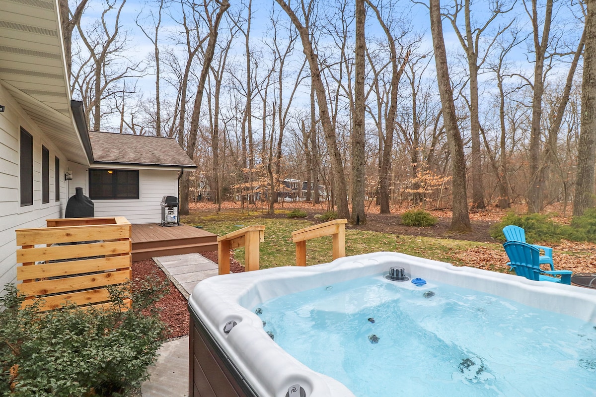 A hot tub is set on an outdoor deck, surrounded by a wooded landscape. The trees are bare, indicating a seasonal change. A lawn area is visible nearby, along with wooden steps leading towards the house, enhancing the connection to nature.