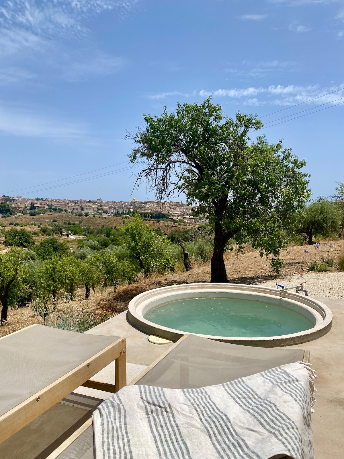 An outdoor area features a round, unmonitored tub filled with water, surrounded by a spacious sunbathing area with lounge chairs. Olive trees and a scenic landscape are visible in the background, with a distant view of a town under a clear blue sky.