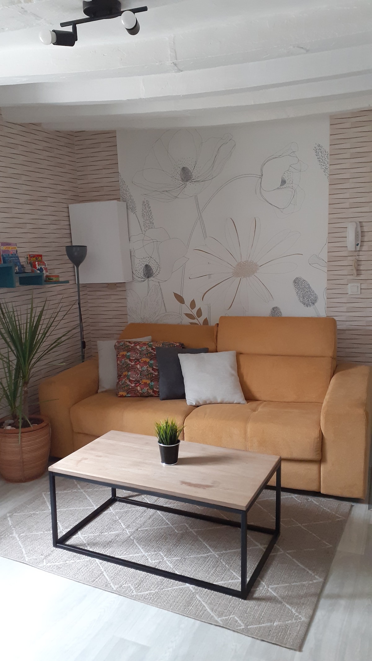 A comfortable living area features a yellow sofa adorned with decorative cushions. A minimalist coffee table rests on a textured rug, complemented by a small plant. Soft lighting is provided by a standing lamp, and a floral wall design adds a touch of elegance.