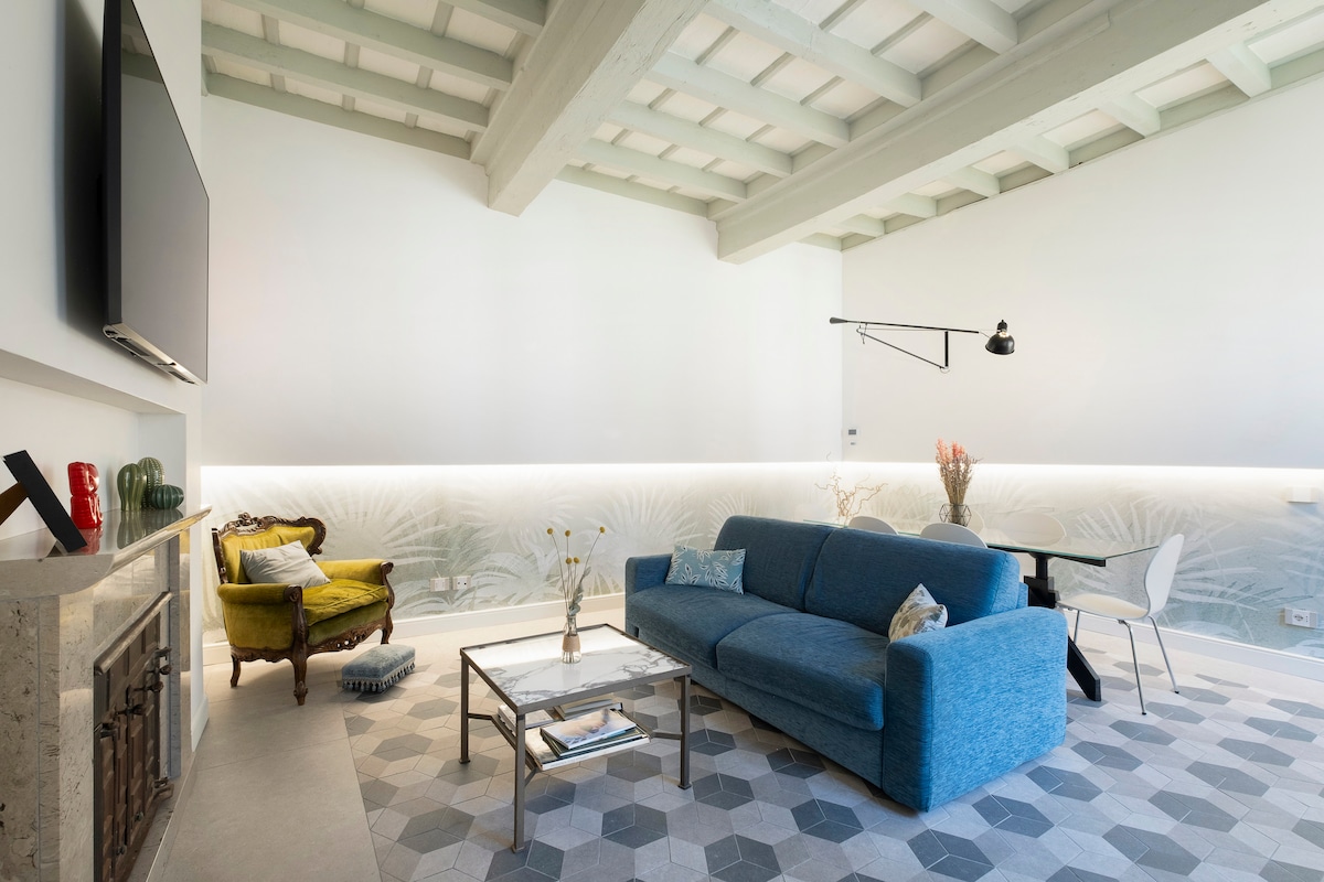 A bright living area features a blue sofa, complemented by a green accent chair. A coffee table with decorative items rests on a geometric patterned rug. A mounted television is positioned above a minimalistic fireplace, while soft lighting enhances the room's modern decor.