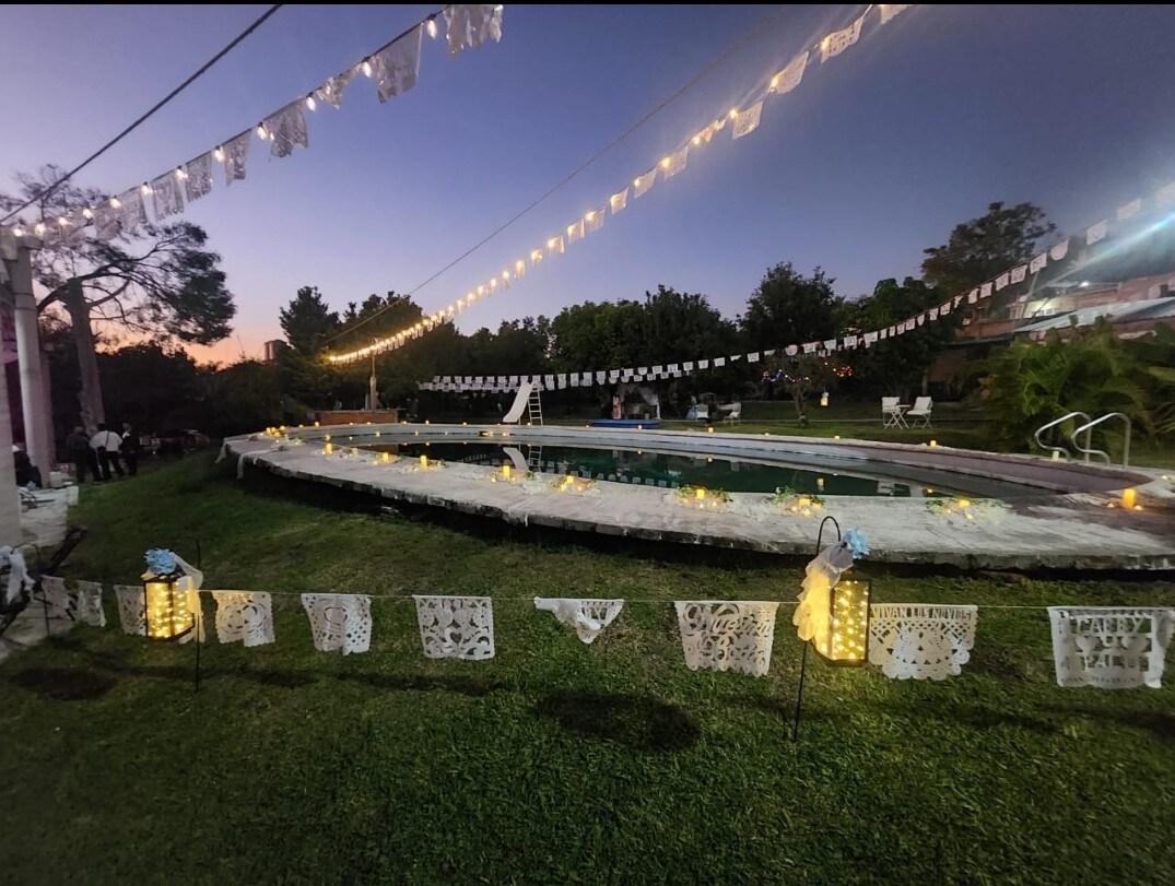 A serene outdoor area is illuminated by string lights at dusk, featuring a winding pool surrounded by lush greenery. Decorative lanterns are placed along a grassy path, with colorful papel picado banners adding a festive touch to the atmosphere.