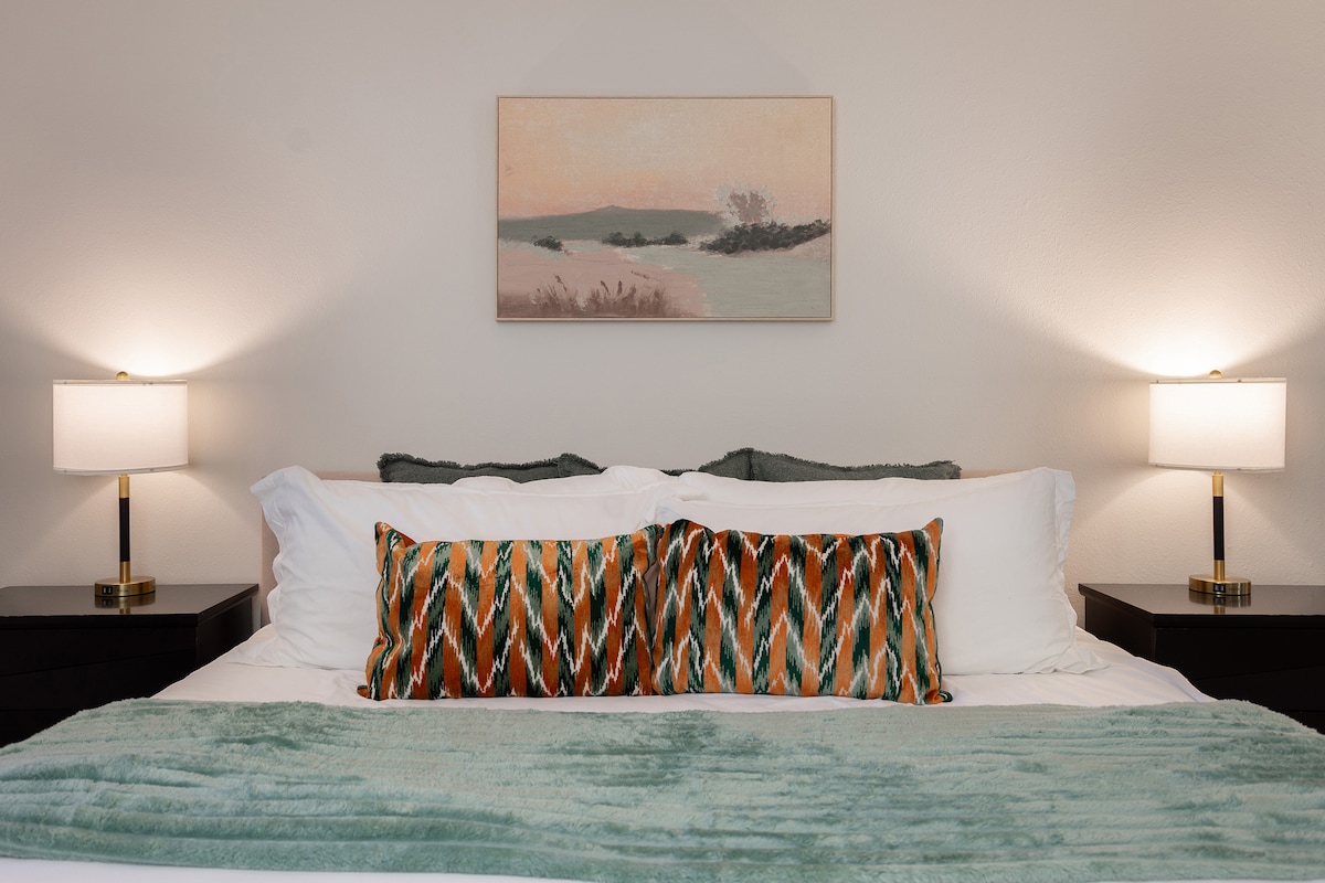 The spacious bed is adorned with a plush blanket and decorative pillows. Two bedside lamps provide soft lighting, complementing a serene landscape painting mounted on the wall above. Nightstands flank each side of the bed, enhancing the overall comfort of the room.