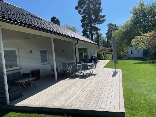 A spacious wooden deck extends from the villa, furnished with outdoor seating and a dining area. A large umbrella provides shade, while a well-maintained lawn stretches out, bordered by trees and greenery, creating a relaxing outdoor space for family gatherings.