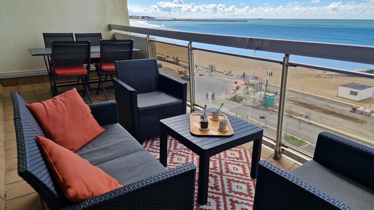 A balcony space features a table with two cups and a small tray, surrounded by four chairs. A patterned rug complements the setting, while plush cushions add comfort to the seating area. The ocean view and sandy beach are visible in the background.