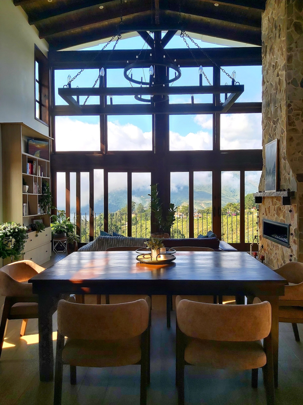 A spacious dining area is featured with a wooden table surrounded by light-colored chairs. Large windows provide expansive views of the mountains and fog, while natural light fills the room, highlighting the stone walls and wooden beams above.