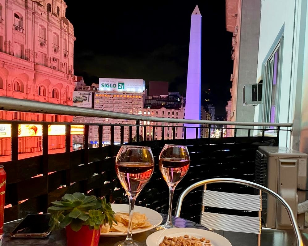 A balcony view features two glasses of rose wine beside plates of snacks. The iconic obelisk is visible in the background, illuminated against the night sky. Surrounding buildings display colorful lights, enhancing the urban ambiance. A small plant adds a touch of greenery to the setting.