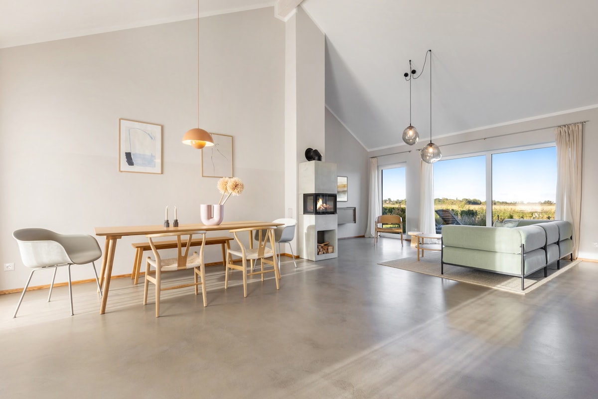 A spacious living area features large glass doors providing natural light and offering views of the surrounding landscape. A sleek dining table is complemented by minimalist chairs. A plush sofa is arranged in a conversational layout near a modern fireplace, adding a cozy touch to the room.