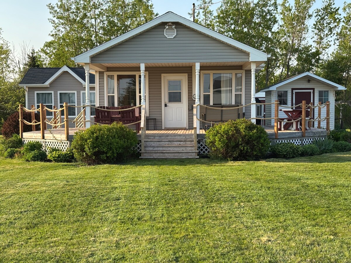 A welcoming cottage with a wrap-around deck is framed by lush green grass and surrounded by trees. The front features a central entry door and two sides with railings, offering a cozy outdoor seating area. Adjacent structures include a guest bunkhouse.