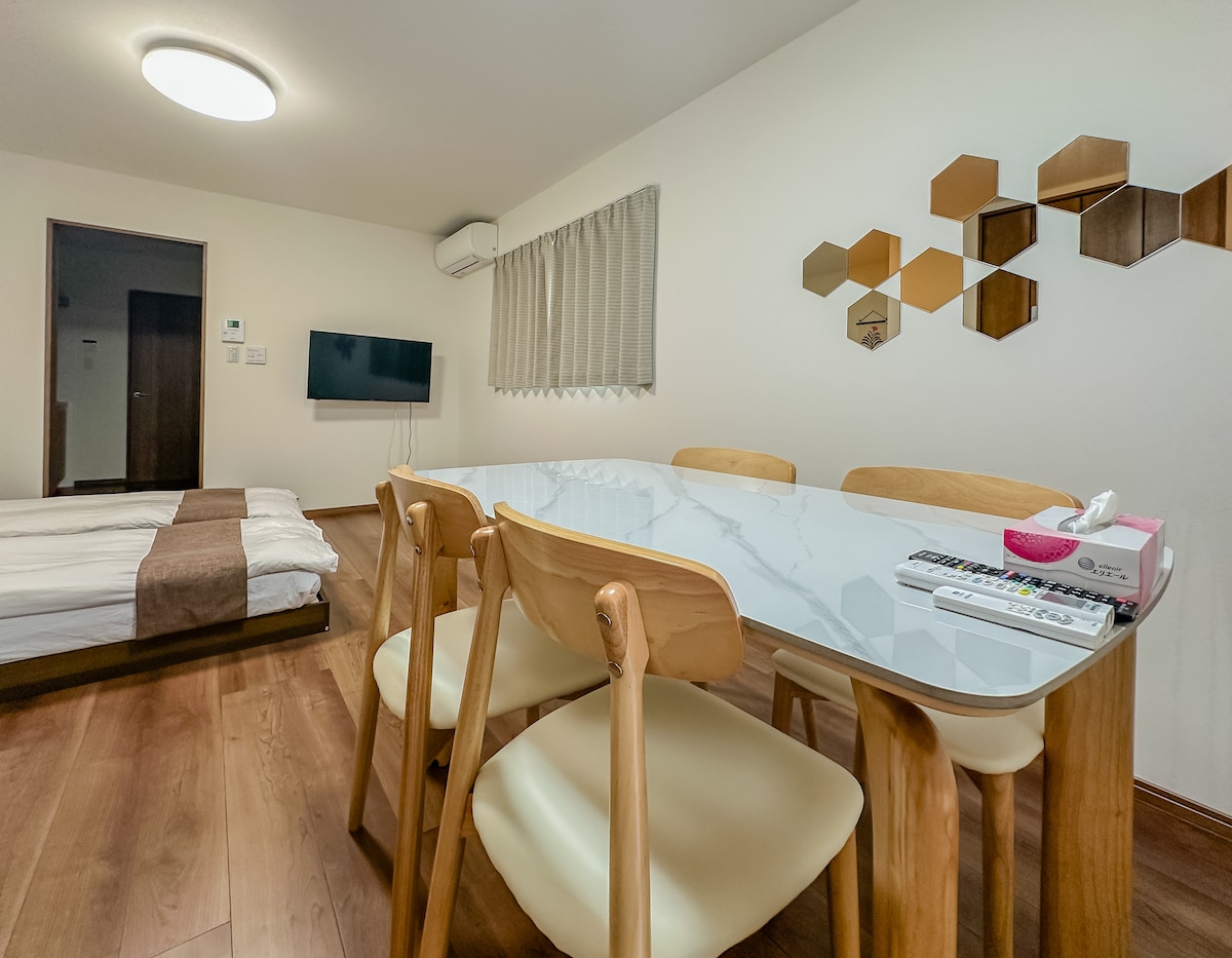 A spacious dining area features a light marble-top table surrounded by four wooden chairs. The room includes a cozy sleeping area with a mattress positioned nearby. Neutral-toned walls are complemented by decorative mirrors, and a television is mounted on the wall.