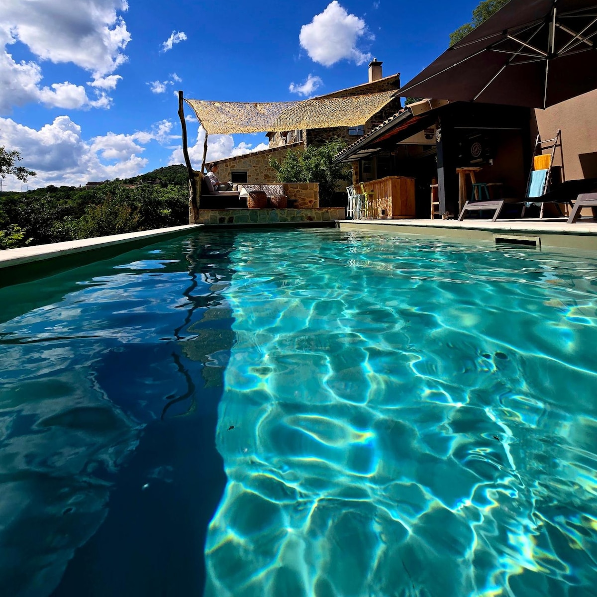 A clear, sparkling salt-water pool reflects the blue sky and fluffy clouds above. Surrounding nature is visible in the background, while a shaded relaxation area is nearby, providing a serene spot to enjoy the outdoor setting.