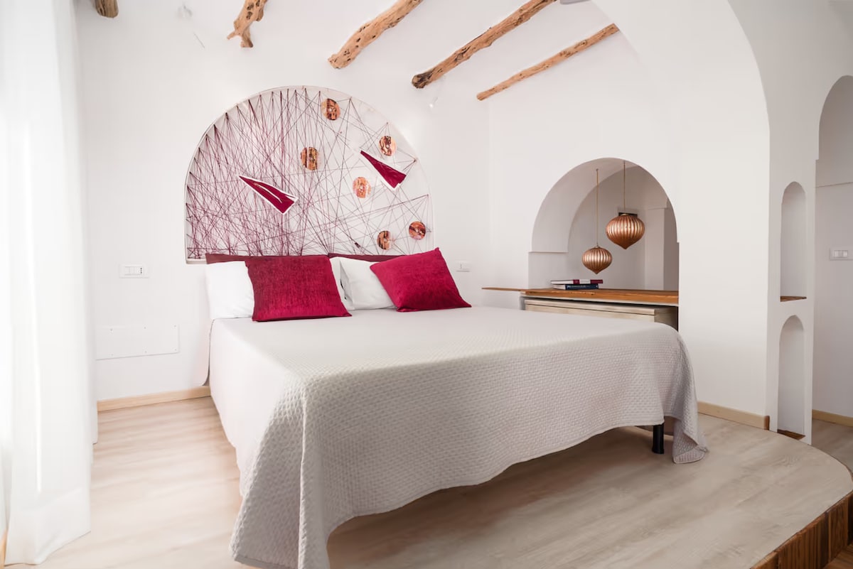 A spacious sleeping area features a large bed dressed in neutral bedding. Decorative red pillows add a touch of color. An artistic wall display is visible above the bed, complemented by wooden beam ceilings and soft natural light filtering through a nearby window.