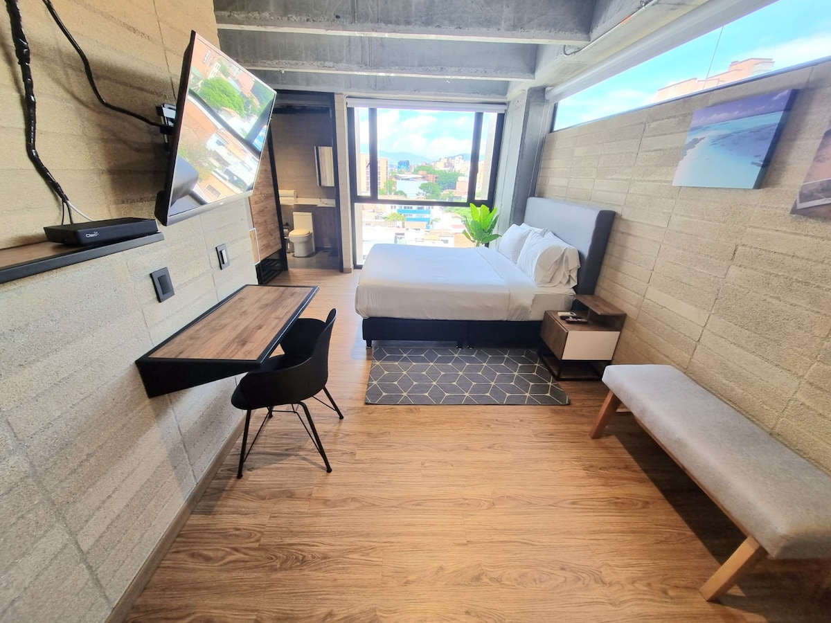 A modern apartment studio is presented with large windows offering scenic views. A spacious bed is accompanied by a desk and chair, providing a comfortable workspace. The calming wooden floor enhances the room's ambiance, while a bench and a television add functionality.