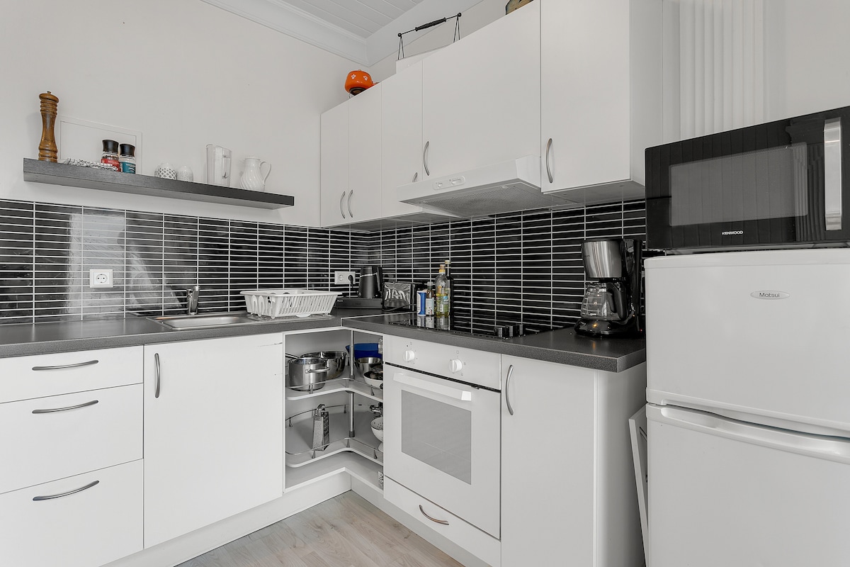 A modern kitchen is equipped with sleek white cabinetry and dark tile backsplash. Essential appliances, including a microwave and refrigerator, are visible. A stovetop and sink are included, with organized storage space and countertop area, offering convenience for preparing meals.