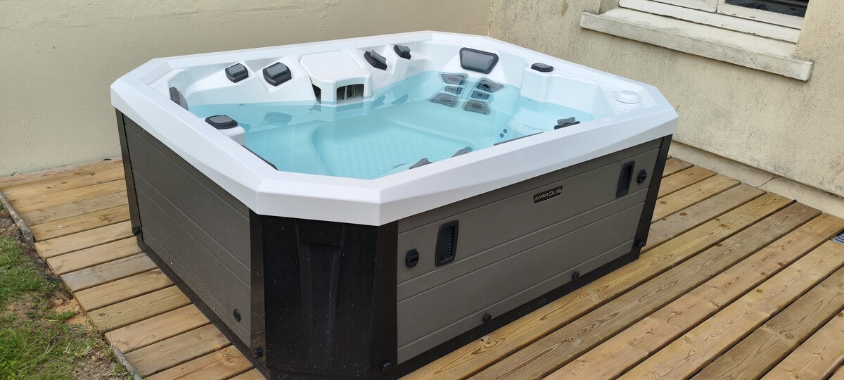 A spacious outdoor jacuzzi, featuring multiple seating areas and jets, is situated on a wooden deck. The water appears calm and inviting, surrounded by a neutral-colored wall, emphasizing the serene setting.