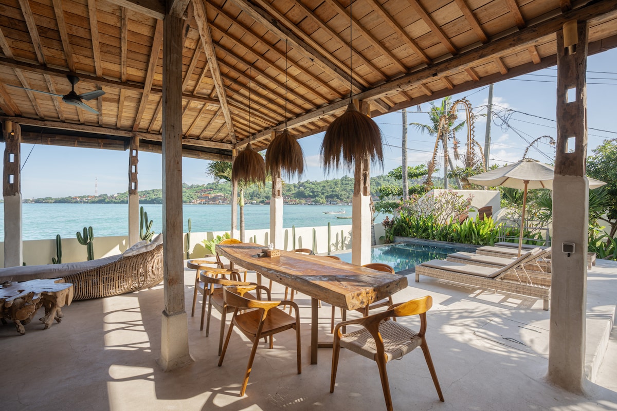 An open living space features a large wooden dining table surrounded by six chairs. The area is illuminated by natural light and showcases a tropical view of the lagoon, with lounge chairs and a pool visible beyond the outdoor seating. Decorative pendant lights hang from the ceiling.
