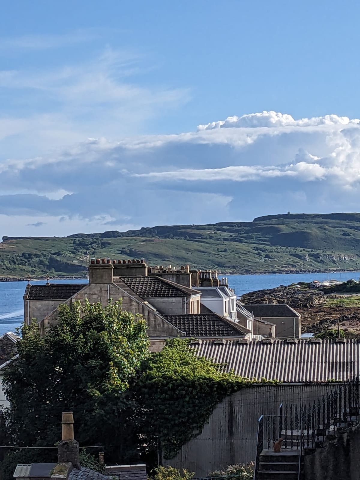 A view of the surrounding landscape, showcasing rooftops and lush greenery with the calm waters in the foreground. Little Cumbrae is visible in the distance, set against a backdrop of dramatic clouds and rolling hills.