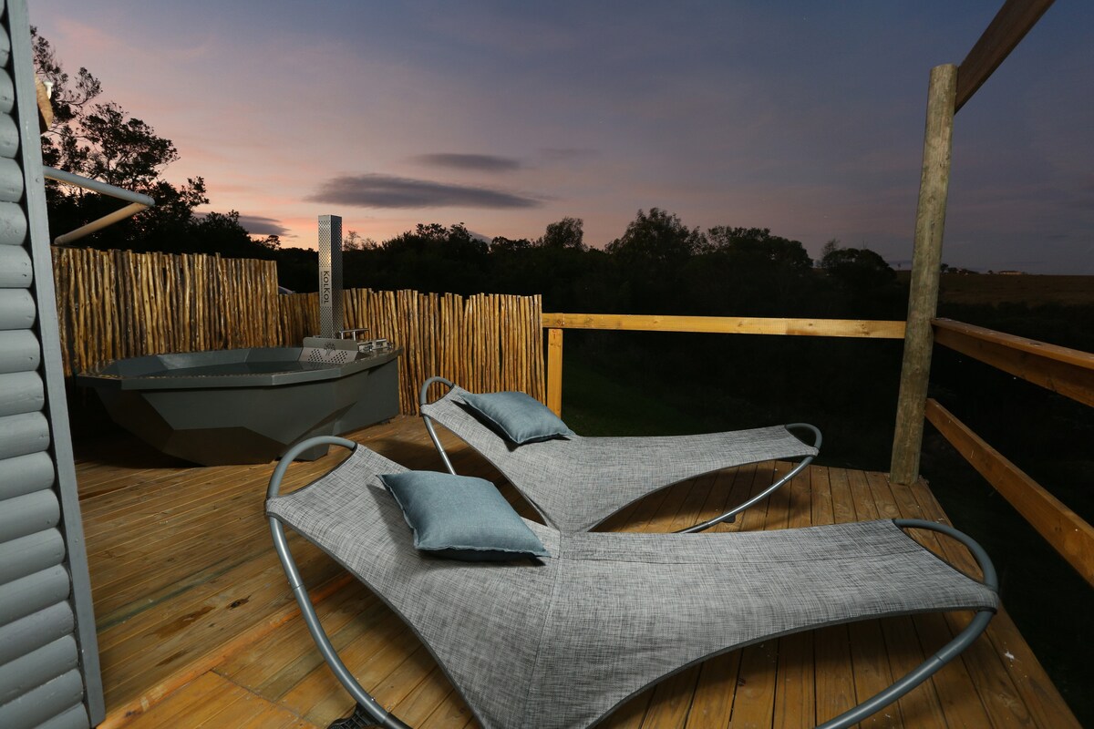 A private deck features two modern loungers with soft cushions, set against a backdrop of a serene twilight sky. A wood-fired jacuzzi is visible nearby, surrounded by natural wood fencing that enhances the peaceful atmosphere.